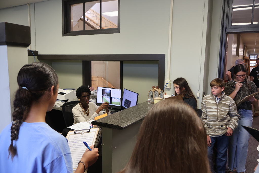 A woman is sitting at her desk with her computer screens beside her while students are standing in front of her listening to her speak. 