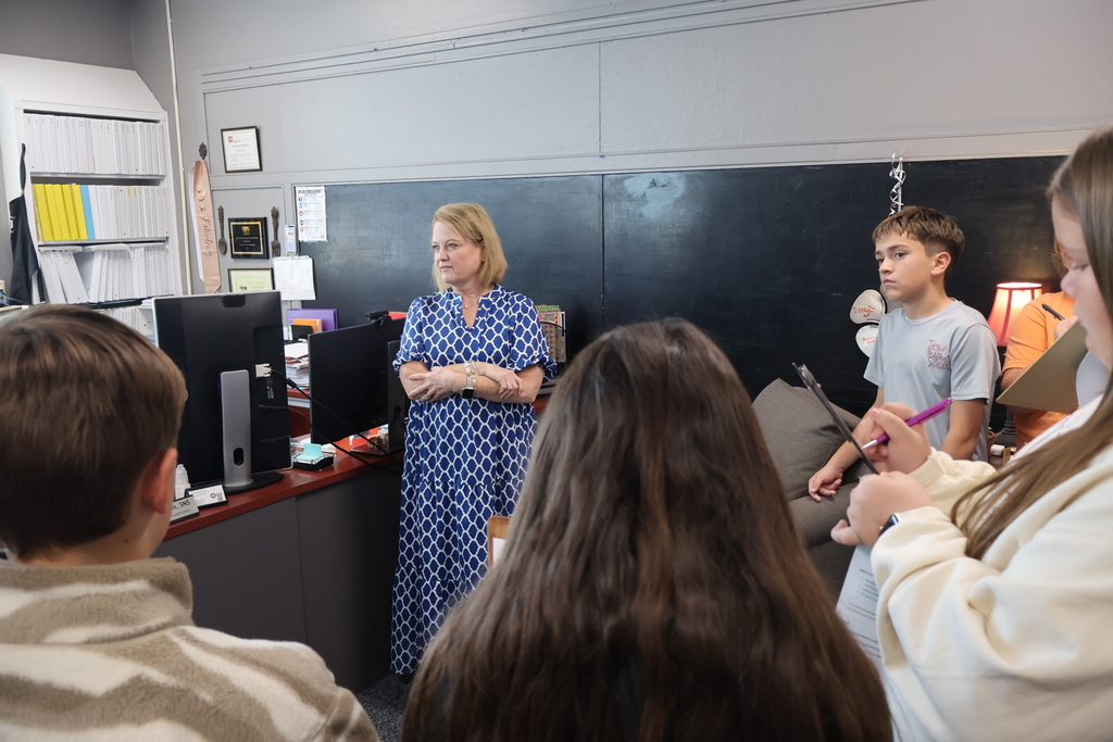 A woman is standing in her office beside her desk while students are standing around her and listening to her speak. Some students are taking notes on clipboards. 
