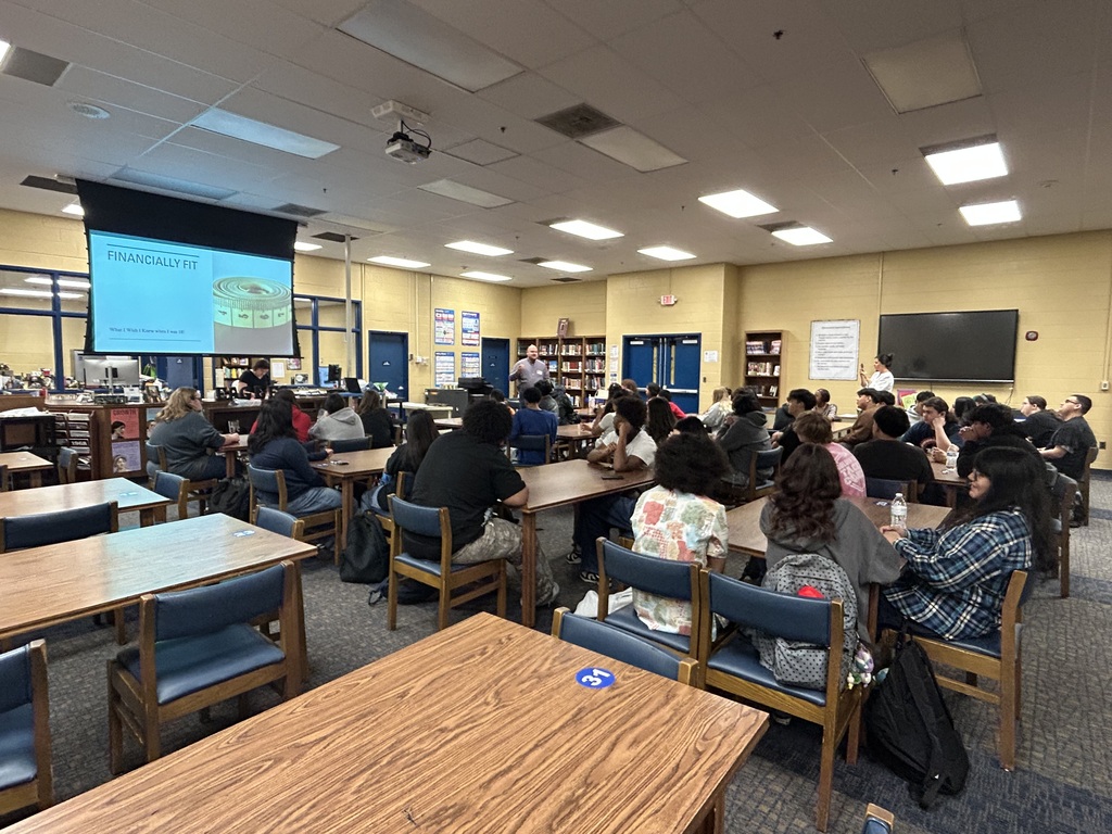 A man is standing in the front of the library speaking to a group of students who are all seated at tables looking at a large screen with a PowerPoint presentation.