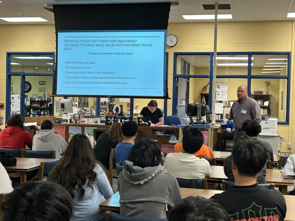 A man is standing in the front of the library speaking to a group of students who are all seated at tables looking at a large screen with a PowerPoint presentation.
