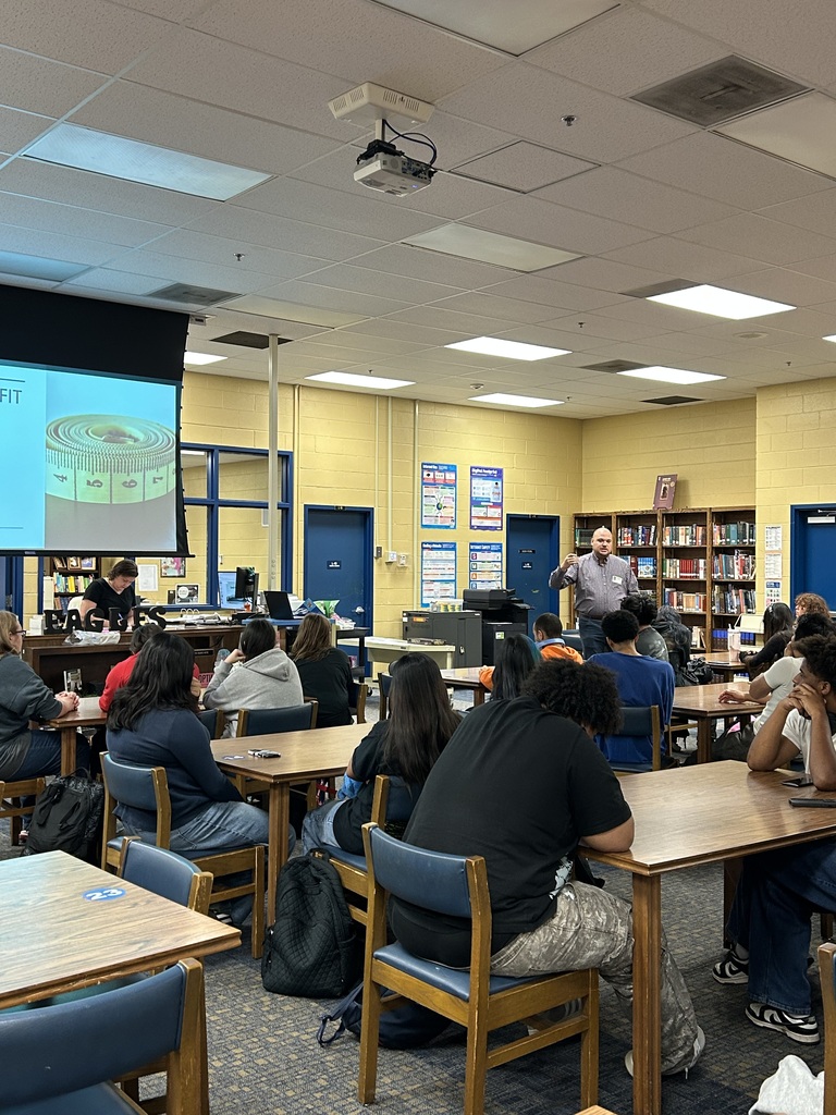 A man is standing in the front of the library speaking to a group of students who are all seated at tables looking at a large screen with a PowerPoint presentation.