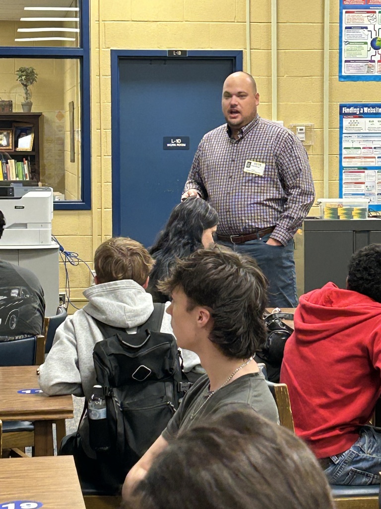 A man is standing in the front of the library speaking to a group of students who are all seated at tables looking at a large screen with a PowerPoint presentation.