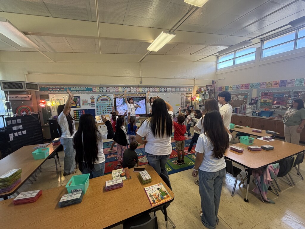 Several high school students are playing "principal says" with the kindergarten students as they all have their hand raised in the air.