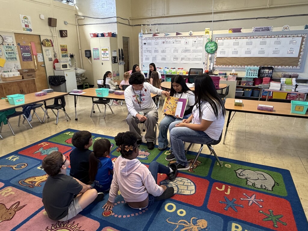 Three high school students are sitting in chairs on the carpet reading a book to four kindergarten students who are sitting in front of them.