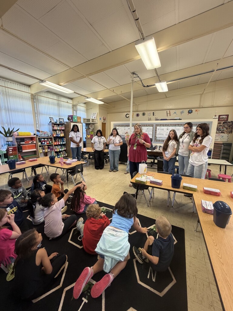 A teacher, along with several high school students, are talking to the kindergarten students.