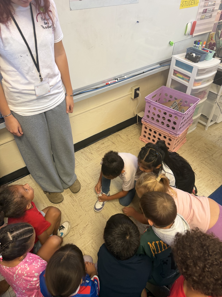 Several kindergarten students are all in a circle learning to tie their shoes from a high school student who is standing in front of them.