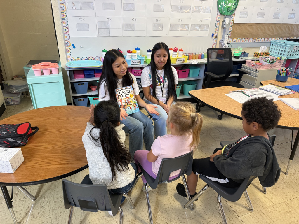 Two high school students are sitting at a table reading a book to kindergarten students who are seated in front of them.