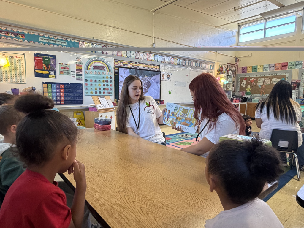 Two high school students are sitting at a table reading a book to kindergarten students.