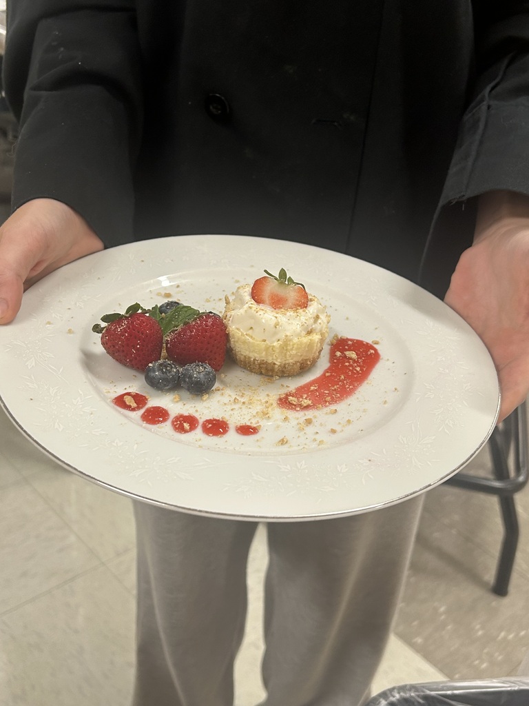 Culinary Arts student in a chef's jacket and hat holds a plated mini cheesecake with berries and red sauce in a classroom kitchen.