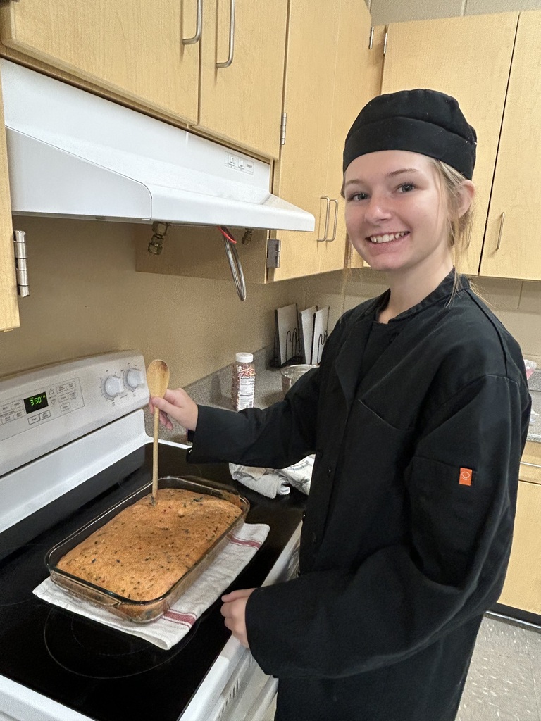 Culinary Arts student in a black chef's uniform and cap holds a wooden spoon over a baked dish in a glass casserole pan beside a stove.