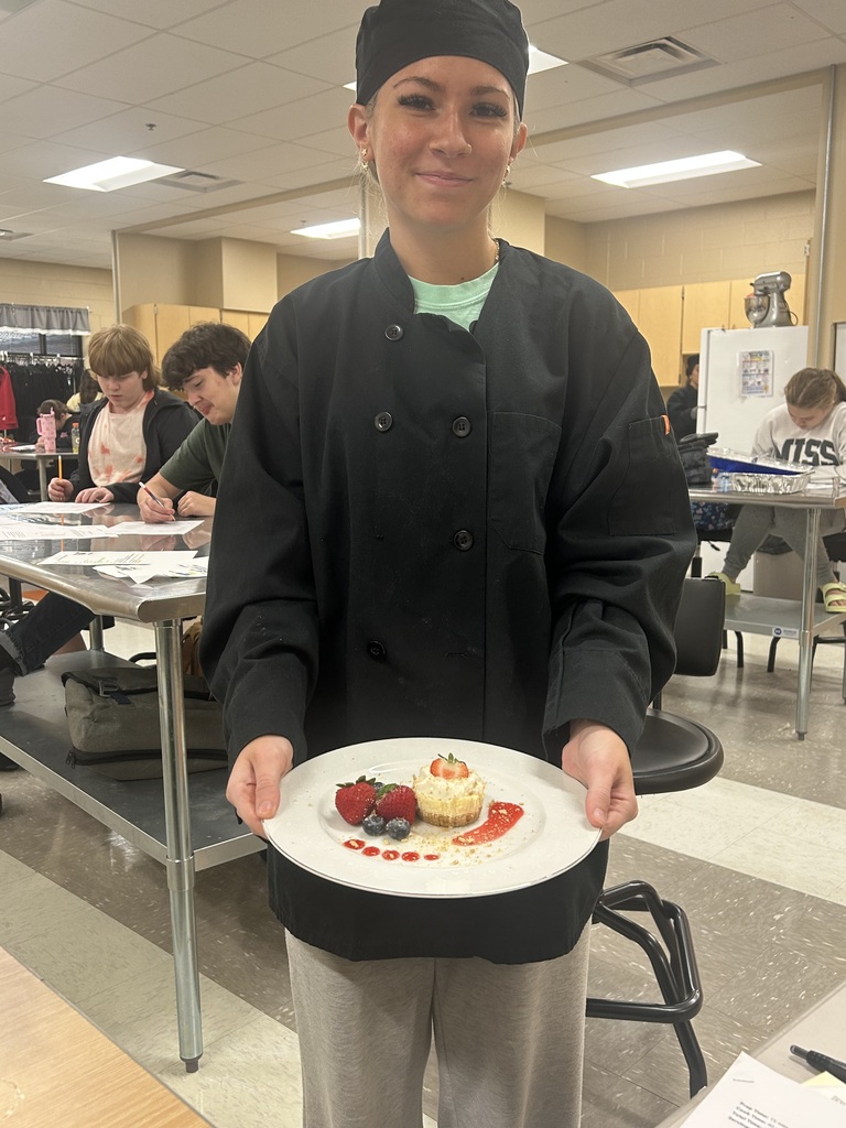 Culinary Arts student in a chef's jacket and hat holds a plated mini cheesecake with berries and red sauce in a classroom kitchen.