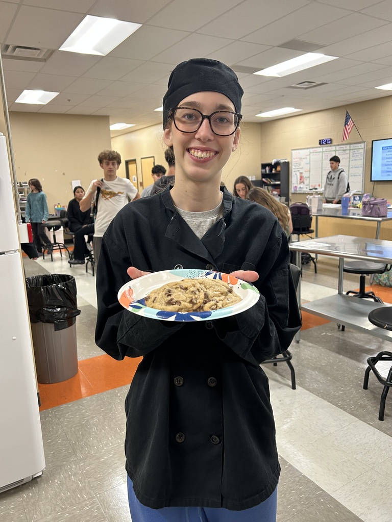 Smiling Culinary Arts student in chef's hat and coat holds a colorful paper plate of chocolate chip cookies.