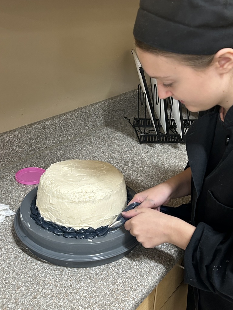 Culinary Arts student in a black chef's uniform smooths the frosting on a round multilayer cake on a turntable