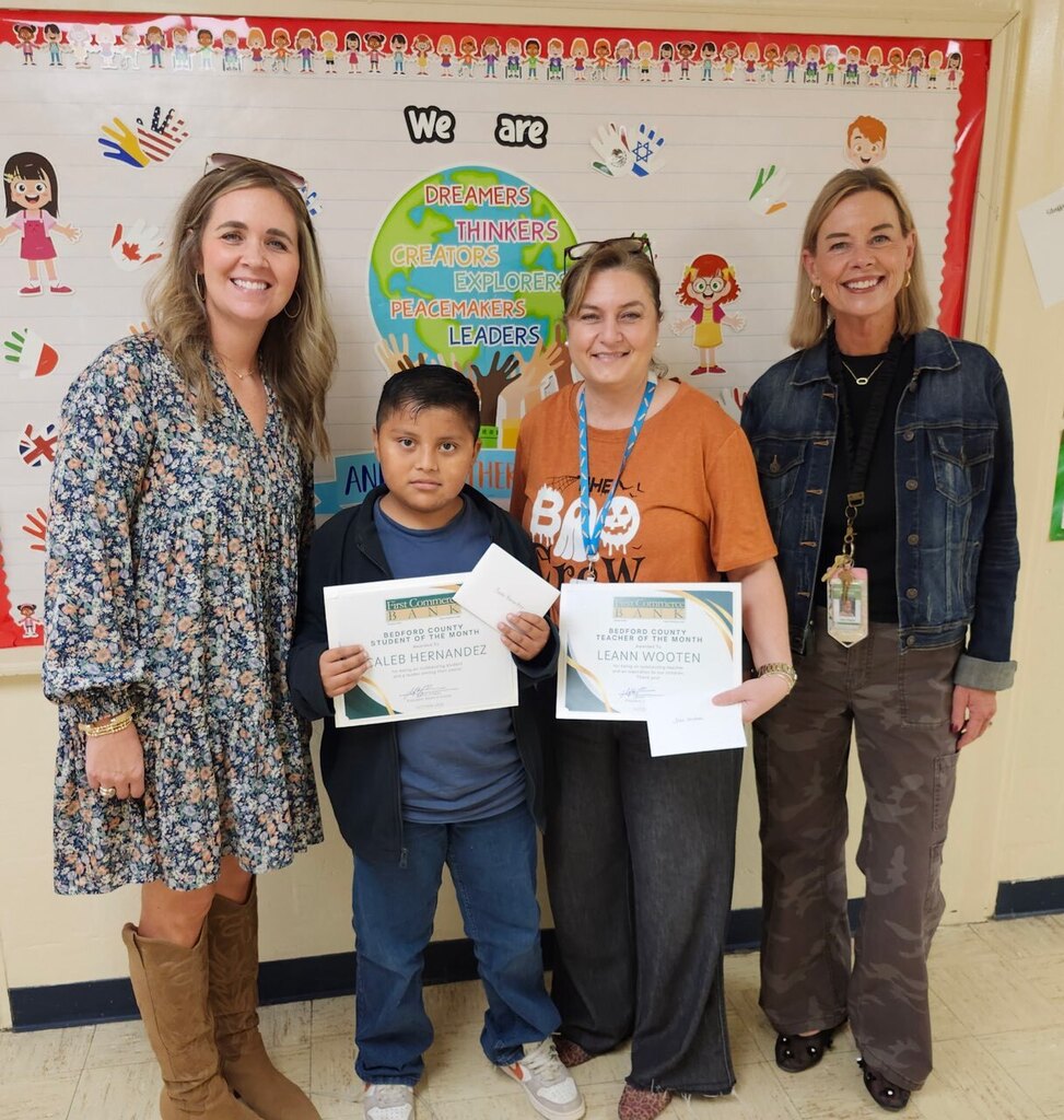 A student and a teacher are standing with two women holding a certificate in the hallway of their school.