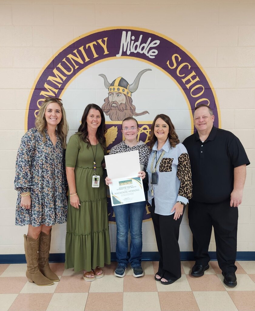 A student is standing with four adults holding a certificate in front of the school's logo that is painted on a wall in the hallway.