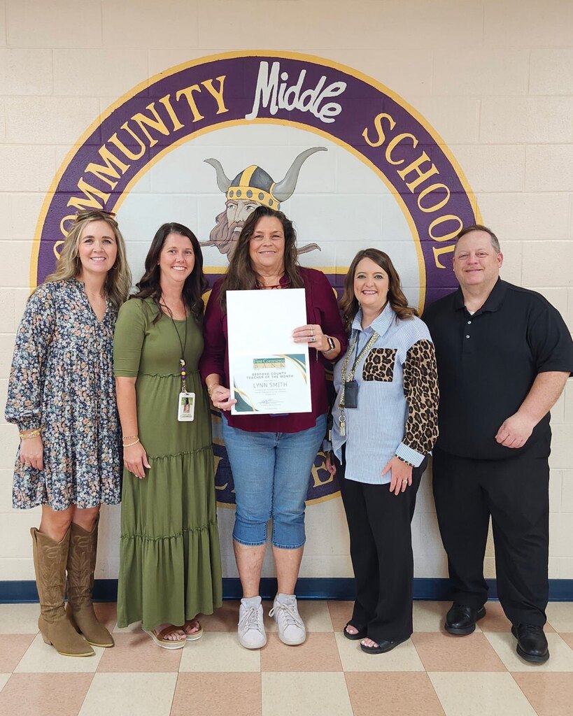 A teacher is standing with four adults holding a certificate in front of the school's logo that is painted on a wall in the hallway.