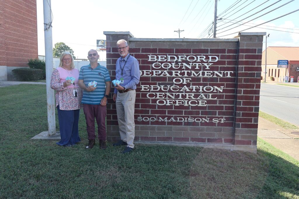 The superintendent along with two others are standing in front of the Central Office sign outside in front of the building and each are holding up three packs of multiplication cards that were donated. 