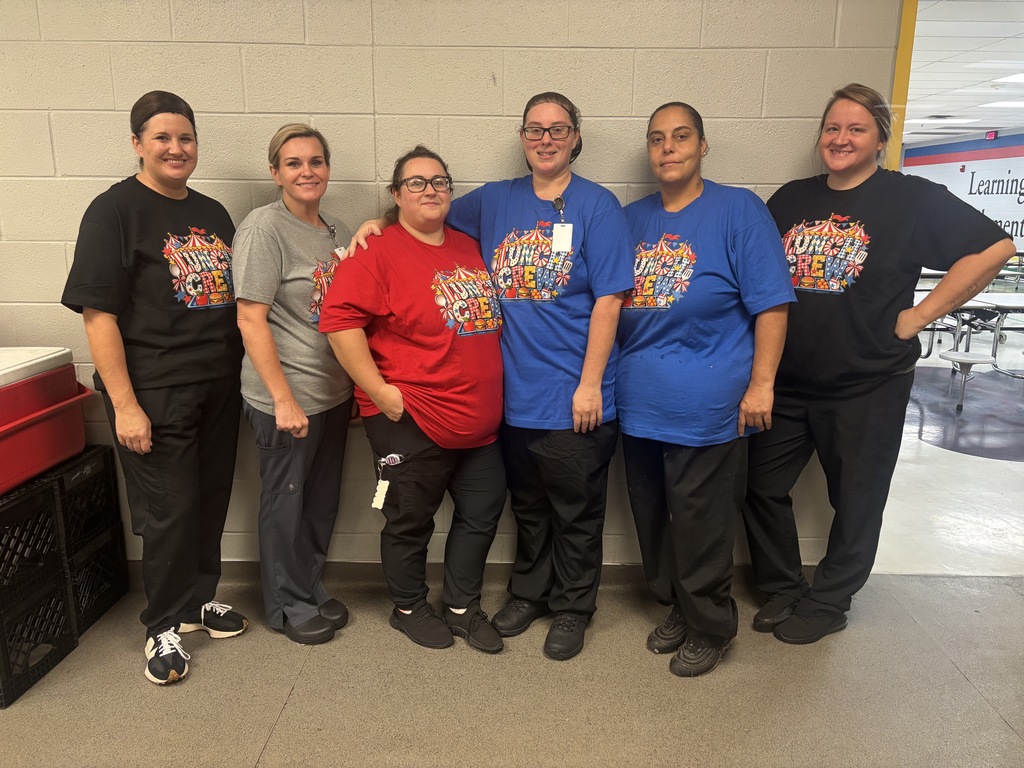 Six women pose for a group picture in their cafeteria dressed in their "Lunch Crew" circus themed matching shirts. 
