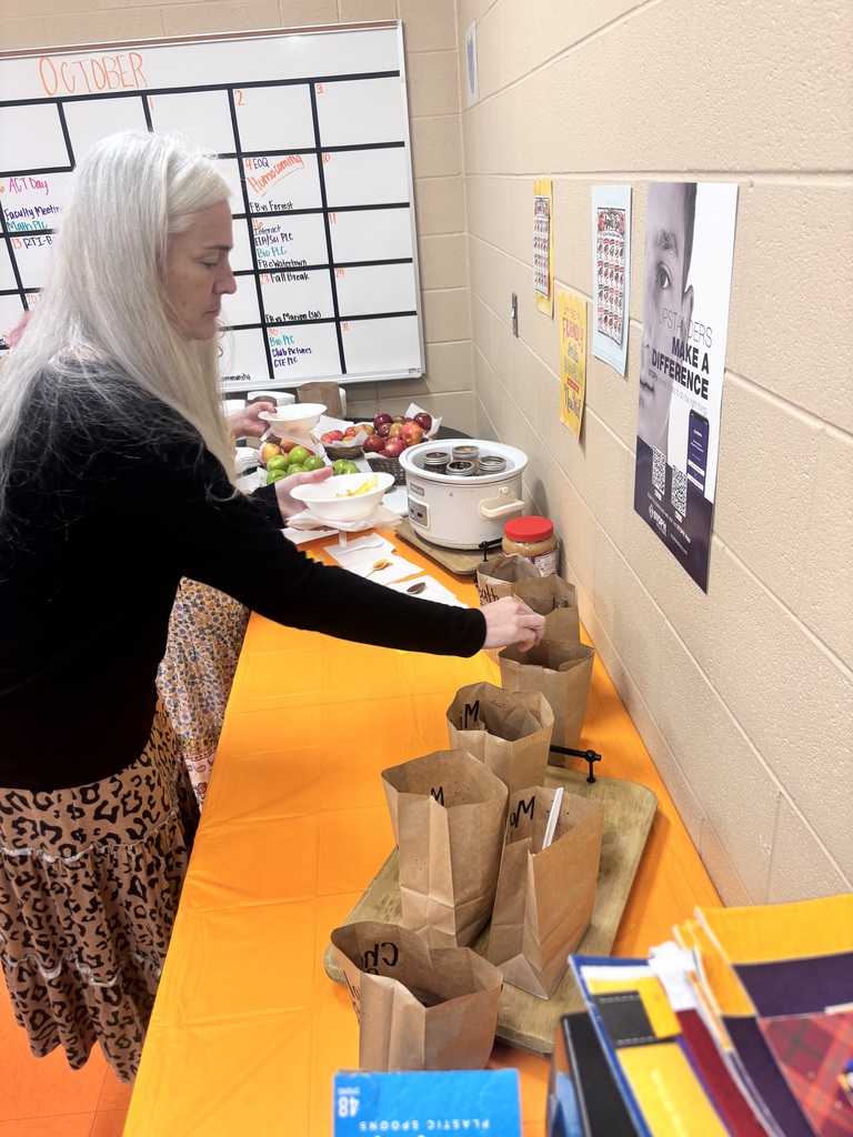 Staff member serves food at an orange-covered table with bowls of fruit and a food warmer, with an October calendar on the wall behind her.