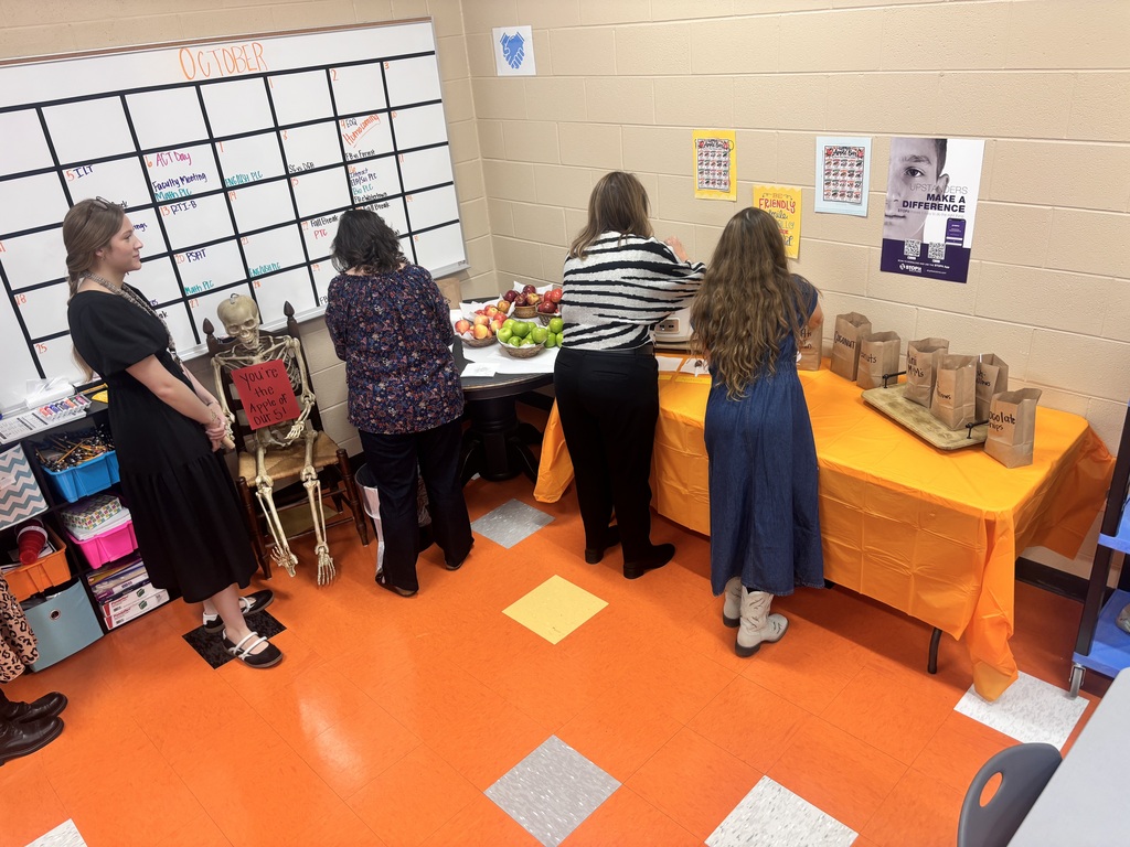 Staff gather around tables with fruit and bags of toppings for an apple bar, with a skeleton holding an apple-themed sign nearby