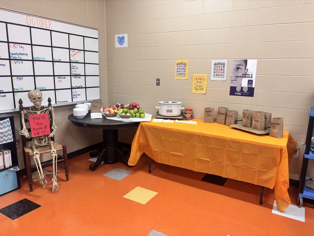 Classroom corner decorated for October with a skeleton holding an apple-themed sign beside a table of apples, fruit, and topping bags.