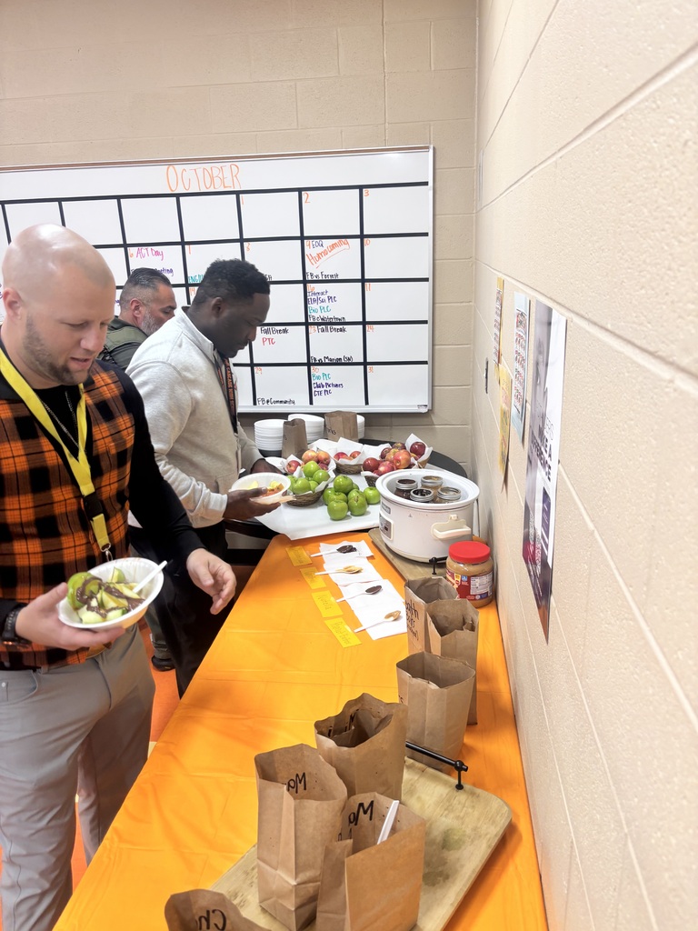 Staff gather around tables with fruit and bags of toppings for an apple bar, with a skeleton holding an apple-themed sign nearby