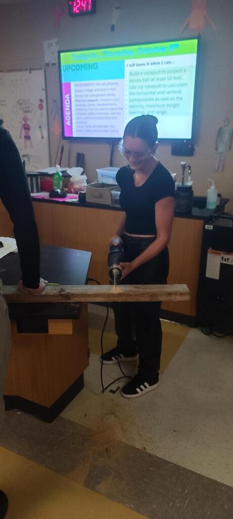 Physics student drills into a long wooden board while a classmate holds it steady, with the lesson goals displayed behind them.