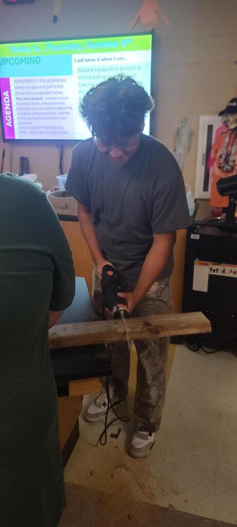Physics student drills into a long wooden board while a classmate holds it steady, with the lesson goals displayed behind them.