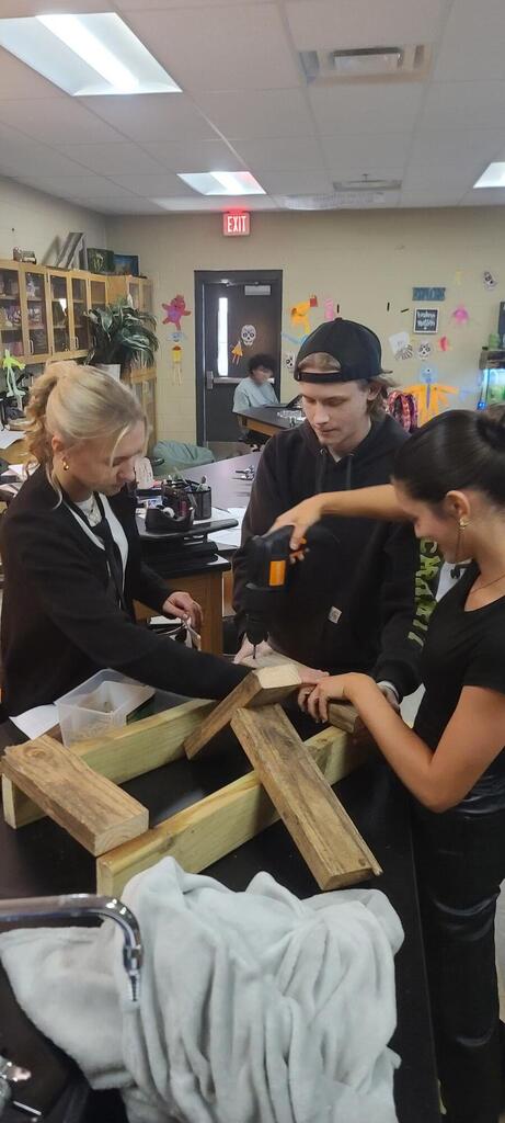 Three students gather around a long wooden bar, one drilling and another steadying it, with a catapult instruction slide behind them.