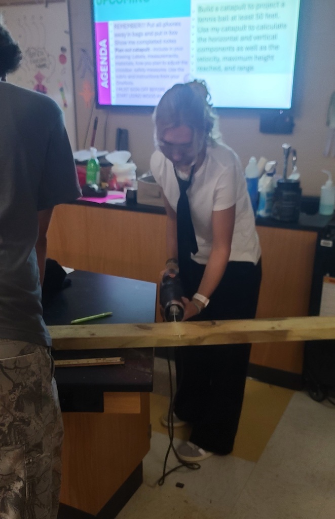 Physics student drills into a long wooden board while a classmate holds it steady, with the lesson goals displayed behind them.