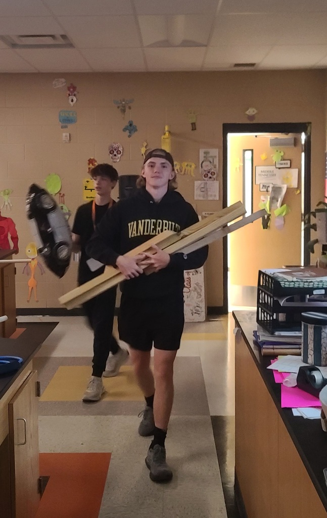 Two students carry long wooden and metal pieces and a large black object through the Physics classroom.