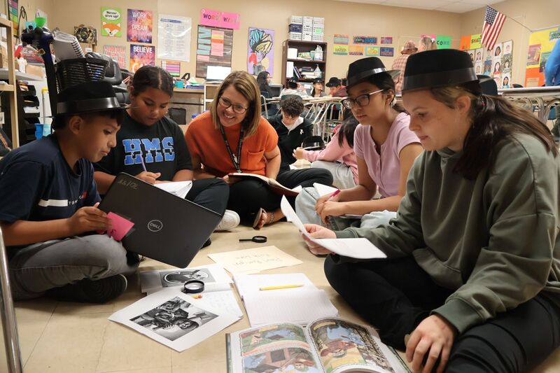 A woman is sitting on the floor with four students while they discuss the book and their findings with her.