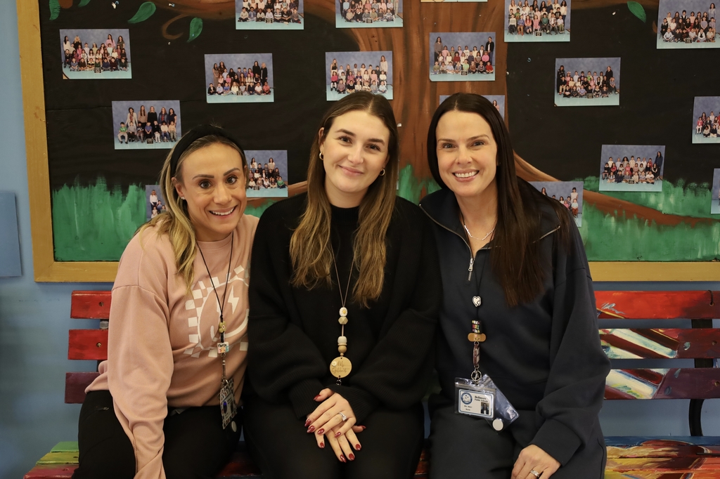 Three women smiling seated on a colorful bench against a mural with tree and photos. They appear friendly and welcoming in casual attire.
