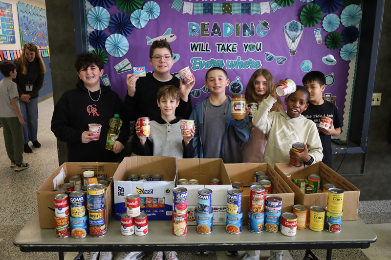 A group of smiling children stand behind a table with canned goods and food items, holding up donations. A bulletin board behind them reads “Reading will take you everywhere.” The scene is cheerful and supportive.