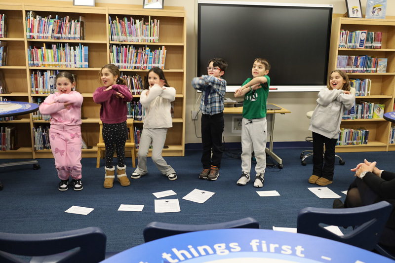 children stand in a library, performing a playful group activity. Laughter and excitement fill the room, with bookshelves and a monitor as the backdrop.