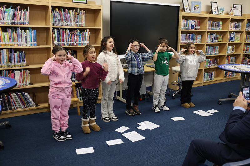 children stand in a library, performing a playful group activity. Laughter and excitement fill the room, with bookshelves and a monitor as the backdrop.