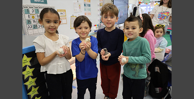 A group of smiling children in a classroom hold certificates and toy chicks, celebrating a "Chick Adoption" day. The atmosphere is joyful and playful.