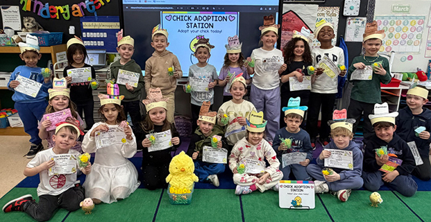 A group of smiling children in a classroom hold certificates and toy chicks, celebrating a "Chick Adoption" day. The atmosphere is joyful and playful.