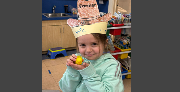 A group of smiling children in a classroom hold certificates and toy chicks, celebrating a "Chick Adoption" day. The atmosphere is joyful and playful.
