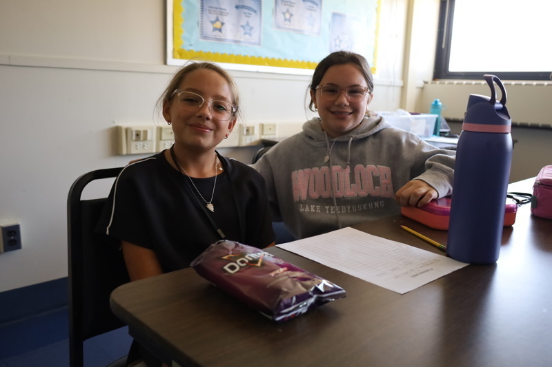 Two people sit at a desk with snacks, water bottles, and a sheet of paper.