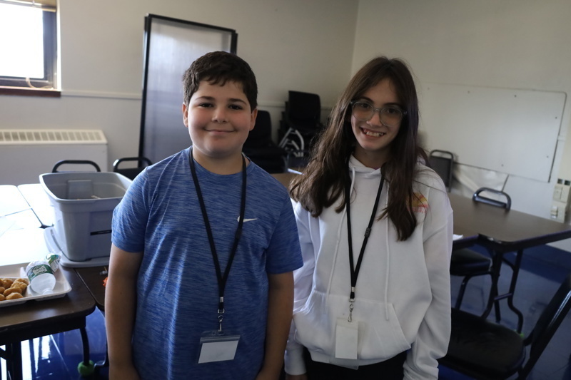 Two people stand in a classroom with tables and chairs, wearing lanyards around their necks.