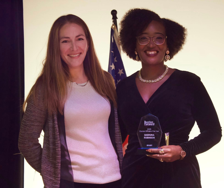 Two women stand smiling on a stage in front of an American flag. Dashuna Robinson, wearing a black dress and glasses, holds a crystal award recognizing her as Elected Official of the Year at the 2025 Berrien Forward Civic Awards.