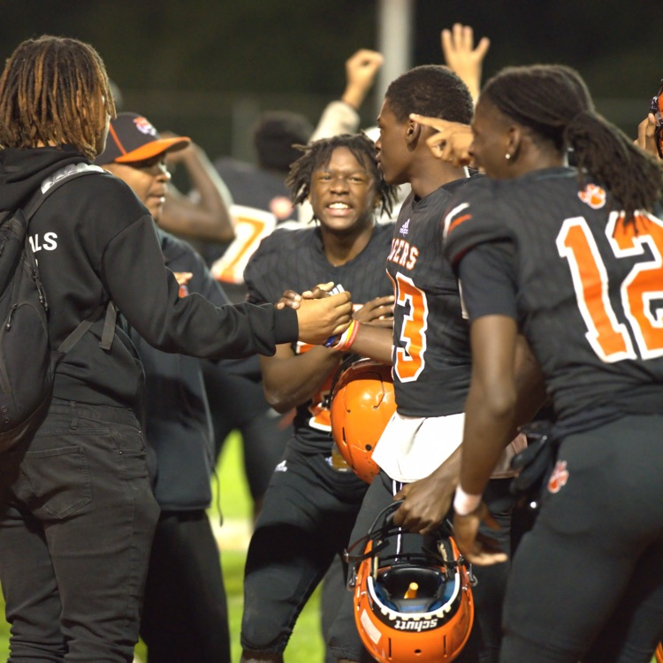 Benton Harbor Tigers football players gather on the sideline, smiling, celebrating, and exchanging handshakes during a nighttime game.