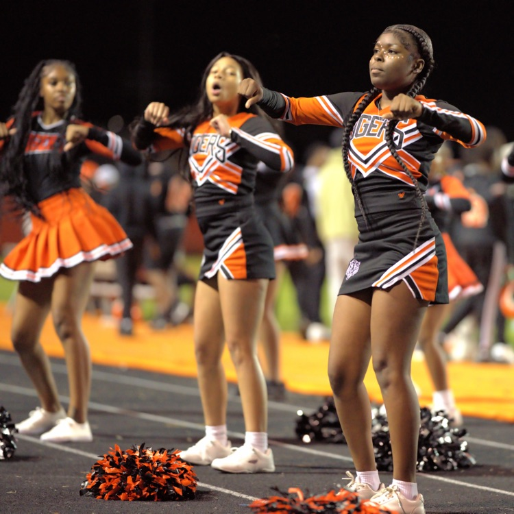 Four Benton Harbor Tigers cheerleaders perform a synchronized routine on the track in orange, black, and white uniforms.