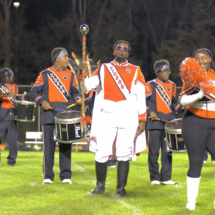 Benton Harbor Tigers marching band percussionists perform on the football field alongside a pom team member waving orange and black pom-poms.