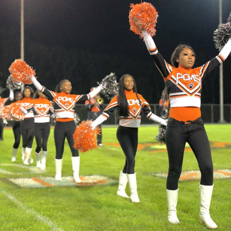 pom team performs on the football field, raising orange and black pom-poms during a nighttime performance.