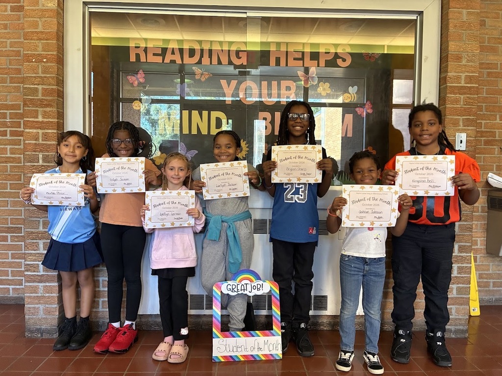 Students standing in front of a bulletin board with the words "reading helps your mind bloom" in big colorful letters. The students are holding their student of the month award certificates.