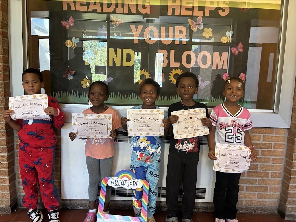 Students standing in front of a bulletin board with the words "reading helps your mind bloom" in big colorful letters. The students are holding their student of the month award certificates.