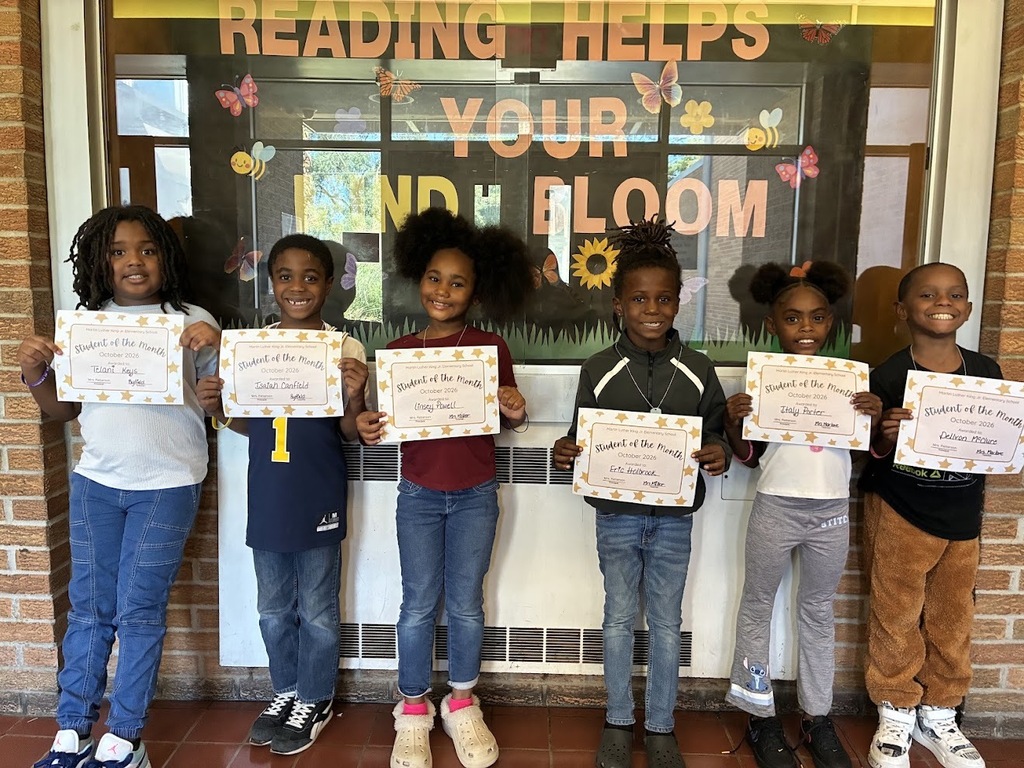 Students standing in front of a bulletin board with the words "reading helps your mind bloom" in big colorful letters. The students are holding their student of the month award certificates.