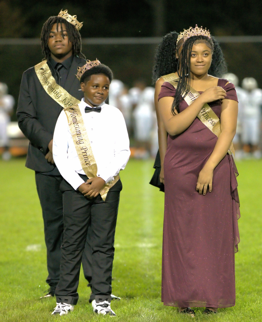 A young homecoming prince wearing a white shirt, black bow tie, and gold sash stands beside a homecoming princess in a burgundy gown and crown.